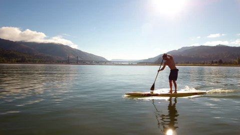Male standup paddler gets a workout while enjoying the view in the Columbia River Gorge.  Aerial shot moving with the paddler on his standup paddle board.