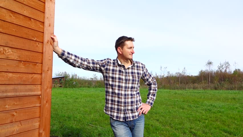 Handsome rustic man stand against wooden house, put hand on hip and look at distance. Slow motion shot, good looking farmer portrait. Rancher wear checkered shirt stand, put hand on timber wall corner