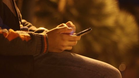 Young boy playing with smartphone in a city park at night. Teenage sitting on bench and touching the phone