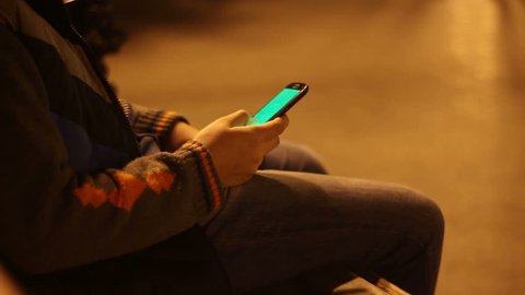 Young boy playing with green screen smartphone in a city park at night. Teenage sitting on bench and touching the phone