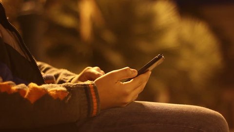 Young boy playing with smartphone in a city park at night. Teenage sitting on bench and touching the phone