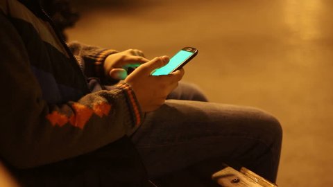 Young boy playing with green screen smartphone in a city park at night. Teenage sitting on bench and touching the phone