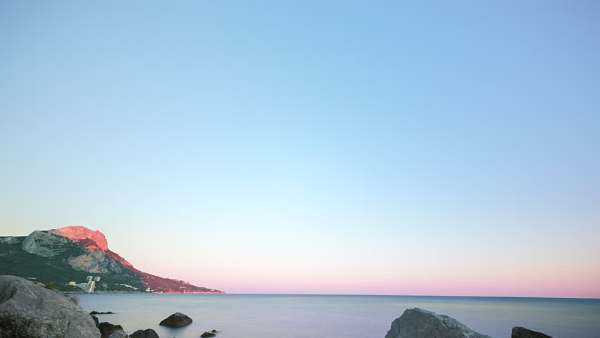 Traces from stars in the form of lines. Cape Sarych is shown as a broad Black Sea coastal landscape with a dark rocky headland, a low mountain silhouette on the left, scattered shoreline rocks in the foreground, and a calm sea stretching to a clean horizontal horizon. The frame begins in deep night with a blue sky filled by fine star points, then the stars extend into long bright trails that sweep diagonally and curve across the upper sky while the coastline remains fixed below. The camera stays completely still throughout the take, holding the same wide composition as the celestial movement builds from short streaks into dense lines and the distant shore lights remain small and steady. As the time-lapse advances, night gives way to blue twilight and then to warm dawn, changing the water from dark gray blue to a pale reflective surface and gradually revealing more detail in the headland. Cape Sarych is associated with a lighthouse that was commissioned in 1898, adding a maritime navigation context to this southern Crimean setting. The final frames show a quiet sea, a fading star field, and a yellow band of sunrise beneath the remaining blue sky, creating a serene nocturnal-to-morning transition.