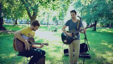 Music band. Duet. Ensemble. Singer and performer. Singer and musician. Two men play a guitar outdoors in summer park. One of the musicians singing into the microphone. Two guitars. Accompaniment.