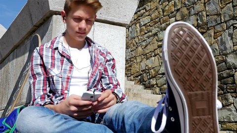 Teenager boy with skateboard and backpack listening to music on smartphone sitting on stairs