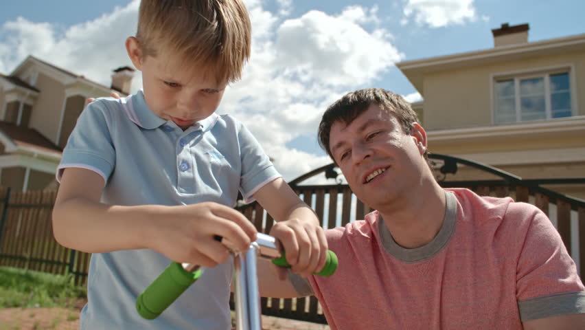 Father happily looking at his little kid fixing handlebar of scooter by himself at summer day