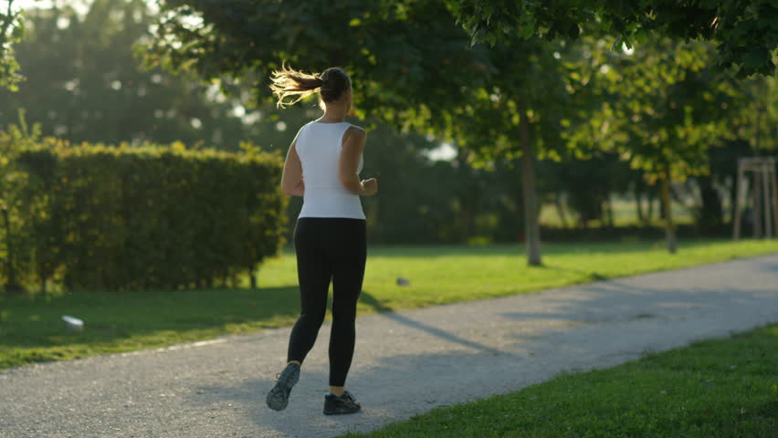 SLOW MOTION CLOSE UP DOF: Attractive young woman in shape jogging and exercising among trees in beautiful park on amazing sunny evening. Tree canopies providing refreshing shade on hot summer day