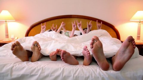 Family makes various gestures by hands in bed under white blanket and then moves barefoot feet
