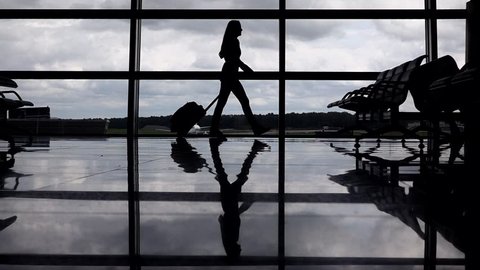Female traveller silhouette, woman go against terminal window with trolley case, slow motion. Empty departure hall, full height passenger, polished floor reflect shadow contour. Cloudy sky outside
