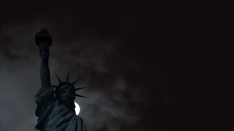 The statue of Liberty, as a dark silhouette, under the pale light of a full moon partially hidden by thick clouds.
