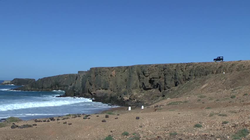 Cliffs at the Atlantic ocean with a classic Landrover on top overseeing the coast. Waves rolling in a small bay at sunny blue skies weather.