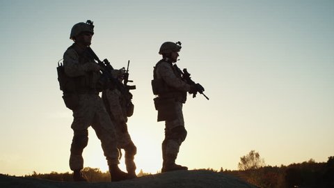 Squad of Three Fully Equipped and Armed Soldiers Standing on Hill in Desert Environment in Sunset Light. Slow Motion. Shot on RED EPIC Cinema Camera in 4K (UHD).