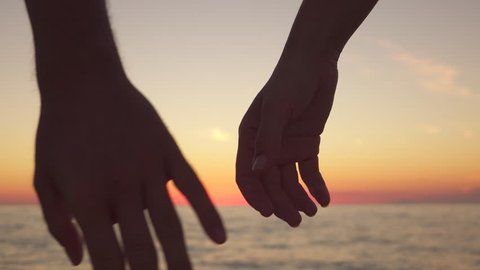 SLOW MOTION, CLOSE UP: Affectionate boyfriend and romantic girlfriend holding hands tightly fingers crossed, showing love, care and intimate friendship to each other at dreamy ocean shore at sunset