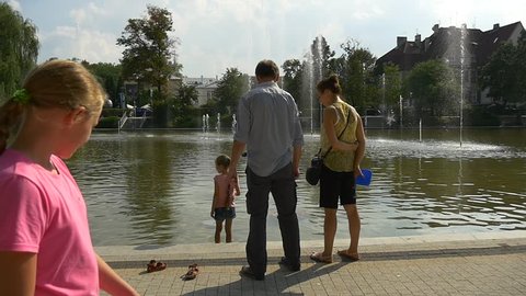 the Little Fair-Haired Girl Stands Knee-Deep in Water. She Looks at Her Reflexion. the Grandfather and Mom of the Girl Speak With Each Other. One More Fair-Haired Girl Passes by Them.