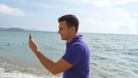 Young smiling man having a video call on smart phone at the sea beach. Happy guy making video online chats on beautiful landscape background. Boy talking on mobile and showing ocean. Man doing selfie