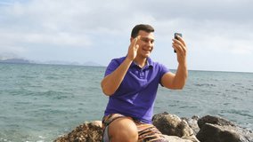 Young smiling man having a video call on smart phone at the sea beach. Happy guy making video online chats on beautiful landscape background. Boy talking on mobile and showing ocean. Man doing selfie
