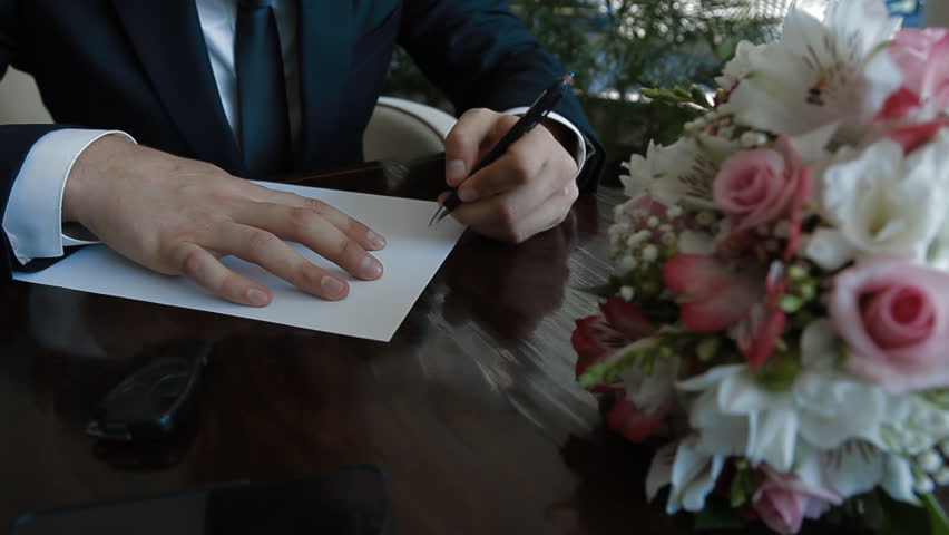 Groom start writing a paper letter to his bride. Weddong bouquet lay in front of man on the table.