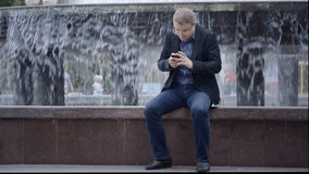 Man Wearing Suit Sitting Near Fountain Writing Sms On His Phone in Slow-Motion 50 fps