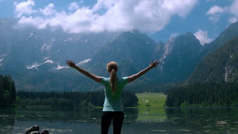 Jib shot - Fit Caucasian female sport fitness model standing near the lake in mountains with arms outstretched toward the beauty of the nature that surrounds her