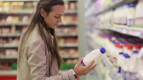 Young caucasian woman in a dairy section of supermarket choosing a bottle of milk. She take a bottle, reading and retuns to the plase, than take one more bottle.
