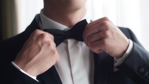Man straightens bow-tie close up slow motion. Well-dressed young man puts and adjusts classic black bowtie on white shirt no face only torso. Success style confidence establishment luxury life concept
