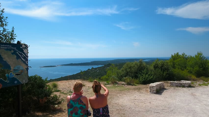 Aerial - Flyover adult daughter pointing her mother on the seascape. Two family members sitting at the island viewpoint and looking at the view