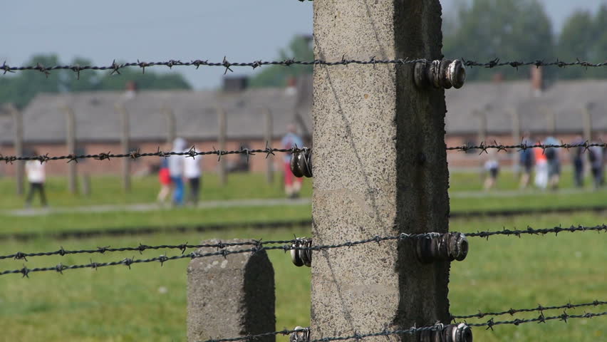Auschwitz: Close up of the fence (detailed), with tourists walking in the background next to the barracks (soft).