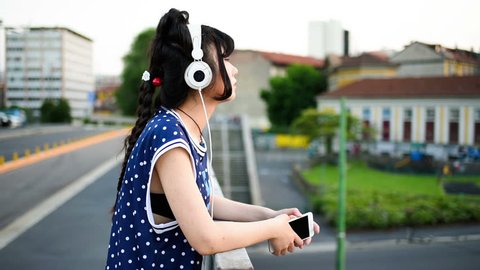 young beautiful asian millennial woman listening music with earphones and smart phone hand hold outdoor in city back light - relaxing, music, technology concept Stockvideo