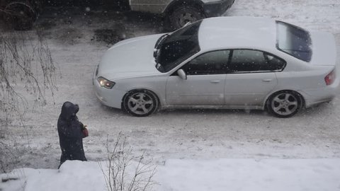 Snow falling in city with a person walking and a car car going on snow covered road in winter. View from above.
