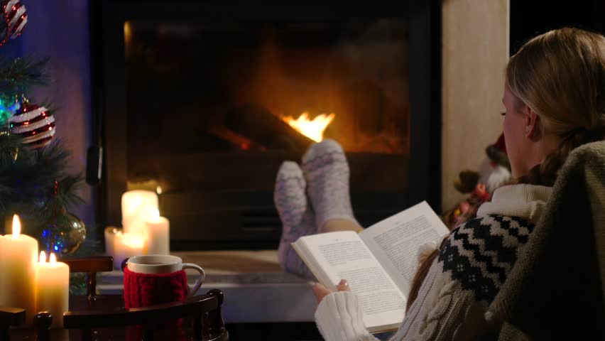 Woman is sitting with cup of hot drink and book near the fireplace
