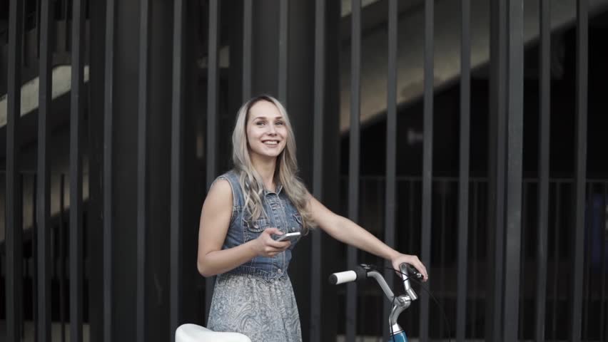 Beautiful smiling caucasian blonde girl standing with a bike using smartphone near grey wall. Lifestyle, technology, communication, urban life, fitness concept.