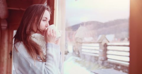 Beautiful Young Woman Looking out the Window and enjoying her Morning Coffee or Tea. 4K SLOW MOTION 120fps. Romantic girl drinking hot beverage at cozy home. Winter Mountains Background. Christmas