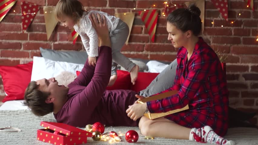 Christmas morning.Young family gleefully congratulates each other Marry Christmas. On bed there are many Christmas gifts in bright wrapping and Christmas-tree decorations. Merry Christmas.
