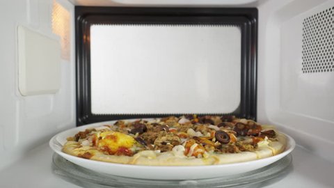 Man reheating baked seafood pizza in the microwave oven. He opens the door of the oven and takes out a dish with hot pizza Frutti di Mare topped with mussels shrimp and olives.