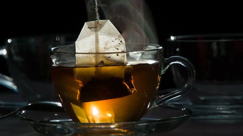 Tea Bag in a Transparent Cup. Teabag falls in a transparent cup with hot water on a dark background.  Water is painted in the color of tea. Shooting at a rate of 240fps.