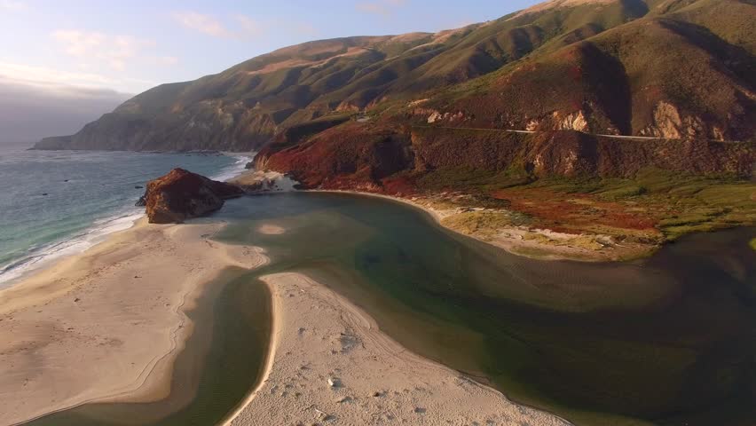 Slow aerial shot moving backwards over the Big Sur coastline in California. View of the Little Sur river emptying into the Pacific Ocean. Sandy beach with green hills and waves crashing. 