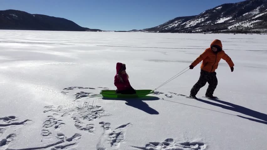 FISH LAKE NATIONAL FOREST, UTAH- Beautiful mountain experience. Winter activities include ice fishing. Two children play on a frozen lake. One child pulls the other on a sled which tips over.