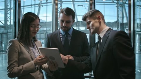 Group of two young Caucasian businessmen and young Asian businesswoman in formalwear using digital tablet and discussing their project in office building elevator