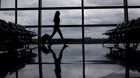 Traveller woman go against lounge window with trolley case, slow motion. Silhouette view, air transportation concept. Empty airport terminal airside area. Self reliant lady proceed to boarding gate