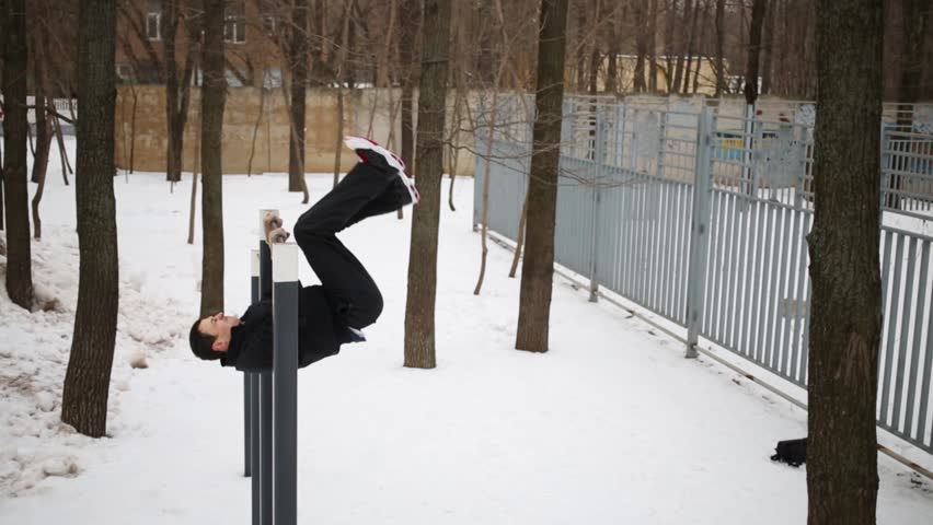 Man in black do hip swing up on horizontal bar and go down at house yard at winter day.