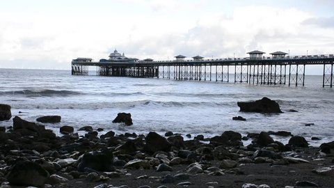 LLandudno pier, seaside resort on North Wales coast, between Bangor and Colwyn bay. Victorian architecture. wide establishing shot with waves and the shore in foreground with rocks.