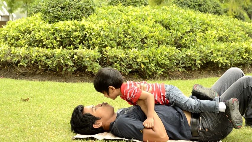 Asian father kisses his son while playing together on the grass in a park.