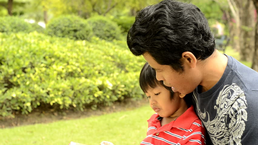 An Asian father and son reading together in a park.
