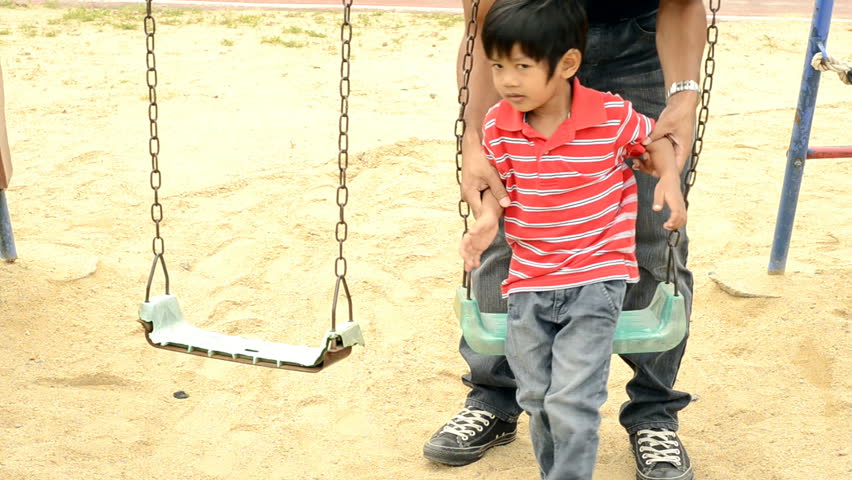 Asian father and son playing together on a swing set, where the boy falls off