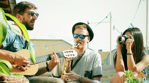 Two charismatic guys playing music with guitar and saxophone at the party while girl taking photos of them and laughing, graded