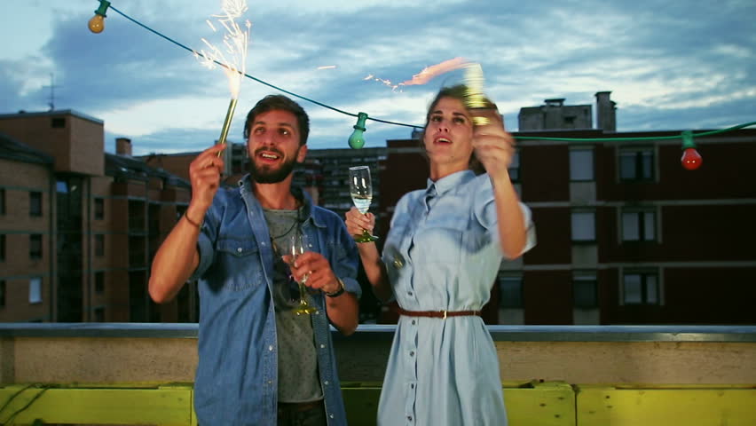 Smiling beautiful couple having fun waving with firework candles and drinking champagne, graded