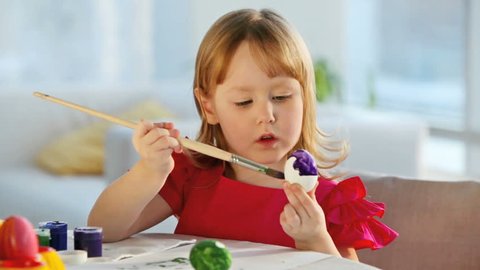 Cute little girl painting eggs with a big brush, pensive and concentrated look
