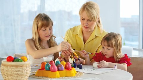 Family of three painting eggs together for Easter, mom explaining the little girl how to make patterns