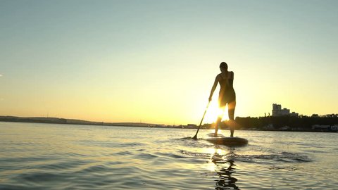 Silkhouette of young woman on Stand Up Paddle Board. Sunset in a city on background. Slow motion. 