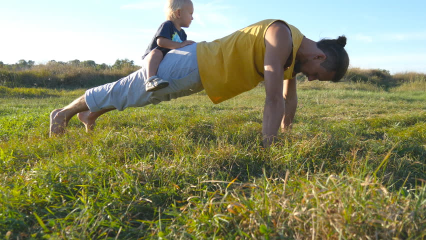 Young muscular father doing push ups with his little son on his back. Athletic man doing push-ups at green grass with happy smiling baby sitting on his rear. Strong guy spending time with your child Royalty-Free Stock Footage #21207259